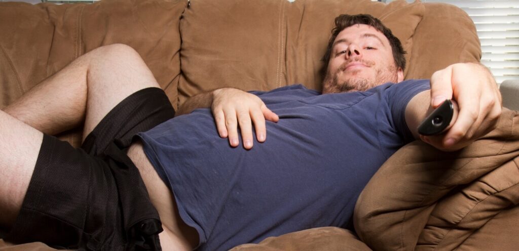 Overweight Man lounging on a brown couch with legs spread, holding a remote control, living a lazy lifestyle