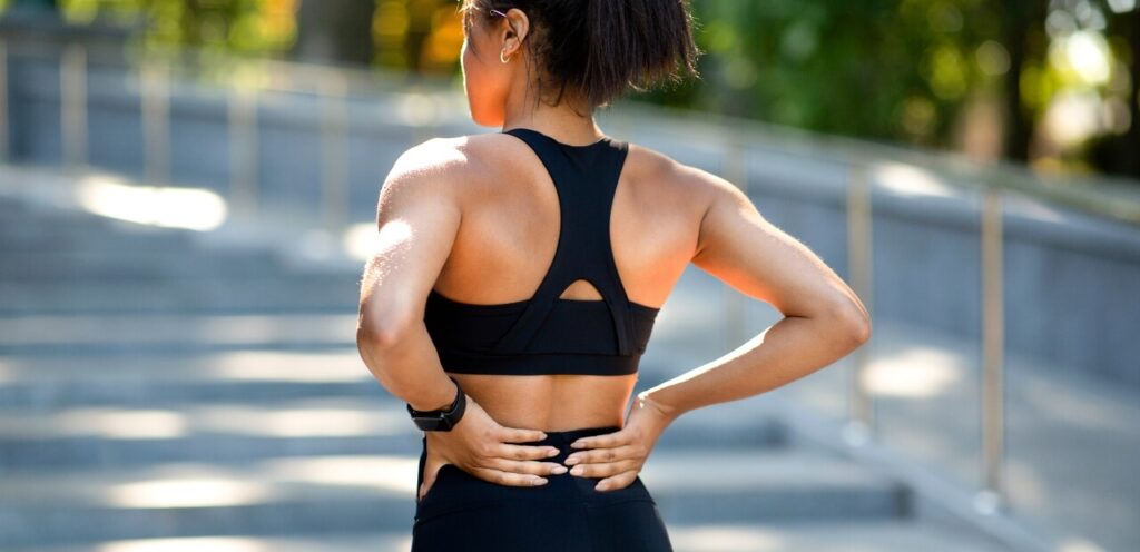 A fit woman in athletic wear, standing outdoors with her hands on her lower back, highlighting the common issue of lower back pain that many face due to various factors like poor posture and ageing