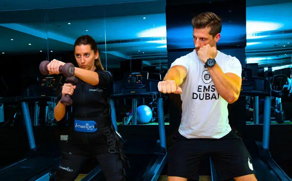 A woman and a man inside a modern gym setting, both in active poses. The woman is wearing an EMS (Electro Muscle Stimulation) training suit branded 'imotion', and is holding hand weights while punching forward.