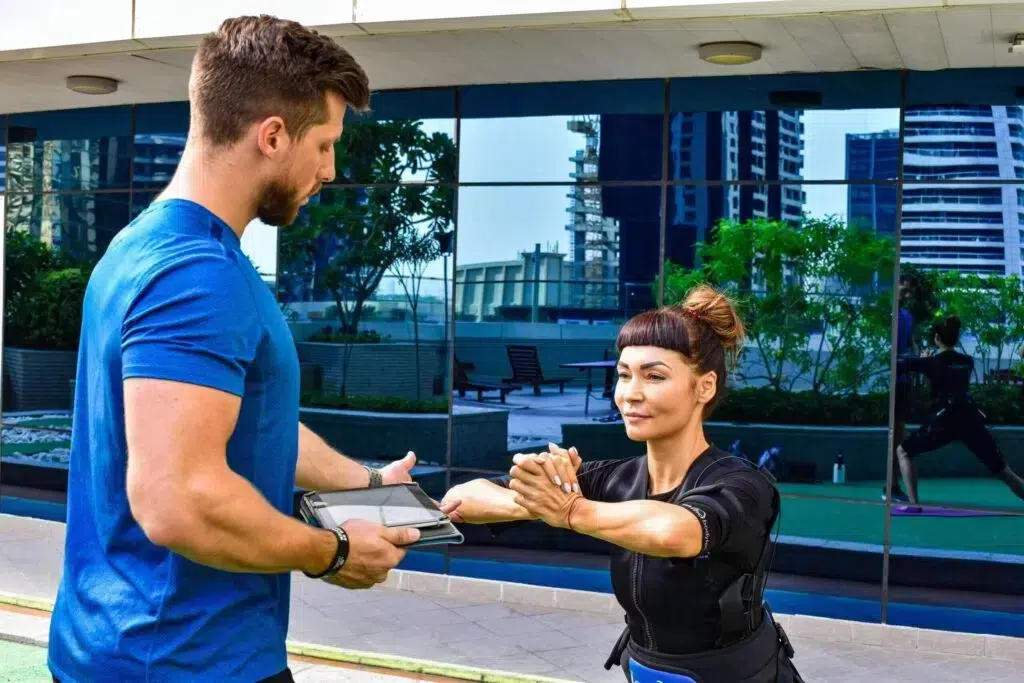 Female participant in an i-Motion EMS suit performs a stretch under the guidance of a male trainer, highlighting the practical application of EMS training in an urban setting.